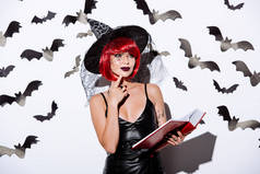 身穿黑魔法万圣节服装、头戴红头发、心事重重的女孩，在白墙边拿着一本印有装饰蝙蝠图案的书