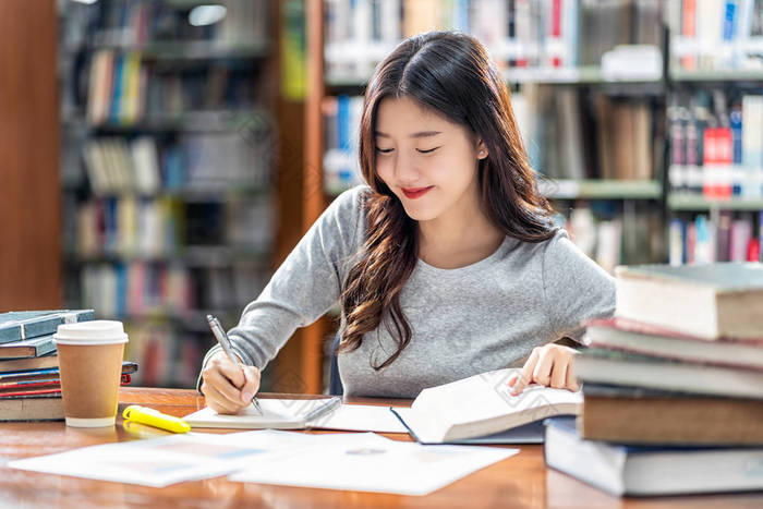 亚洲青年学生穿着休闲服在大学图书馆阅读和做作业，或与各种书籍和固定在书架背景的木桌上，回到学校