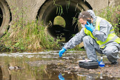 戴防护手套和面罩的男人对河水中的污染物进行快速分析，并用手机传送信息