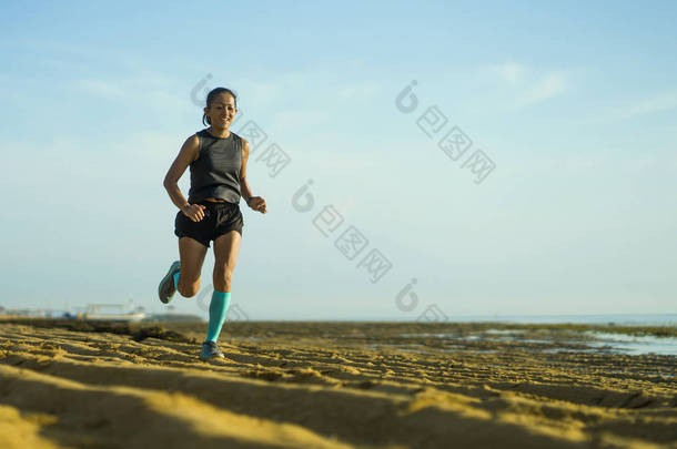 年轻迷人而快乐的亚裔印尼女子穿着压缩袜子在海滩上欢快慢跑做运动的户外健康画像 