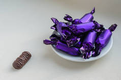 用紫丁香包装纸包在茶托上的成堆的巧克力。白瓷盘盛满了美味的糖果.附近是一个没有包装的糖果。眼睛水平射击。特写。有选择的重点.