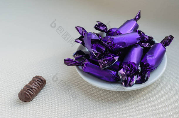 用紫丁香包装纸包在茶托上的成堆的巧克力。白瓷盘盛满了美味的糖果.附近是一个没有包装的糖果。眼睛水平射击。特写。有选择的重点.