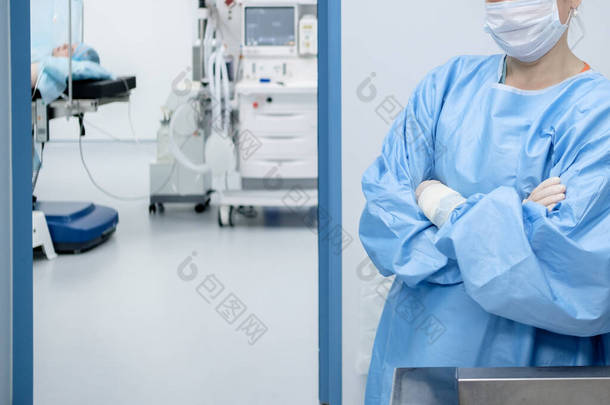 一位穿着手术服、戴着口罩、戴着无菌手套的女外科医生双手交叉地站在手术室入口处。桌子上的手术室里有个病人. 