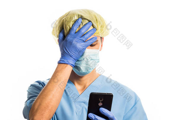 医生身穿医学服装，戴着橡胶手套，戴着面具，一边看着手机，一边用手捂住脸，在白色的背景上拍照