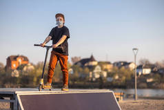 日落时,一个少年戴着蓝色的面罩站在蓝天下.和一辆滑板车一起Covid-19.