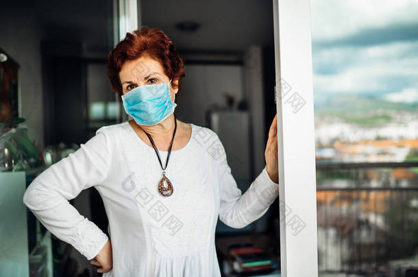 老<strong>年</strong>悲伤的女人在家里，戴着<strong>口罩</strong>，站在阳台的窗户上。考罗纳维勒斯COVID-19疾病爆发感染的危险。
