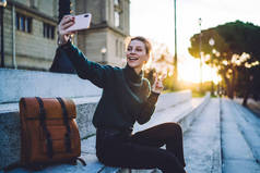 穿着保暖休闲衫、背着背包坐在街上楼梯上的快乐的年轻女子，在朦胧的阳光下，用手机拍下了自己的照片，同时还展示了胜利的标志 