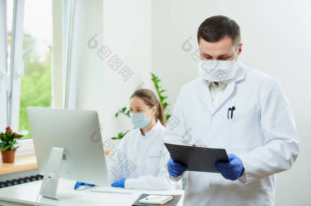 一个戴着假发的外科医生拿着一个黑色的剪贴板，在<strong>医院</strong>里填写病人的<strong>卡片</strong>。一位女医生坐在房间角落里的台式计算机前 