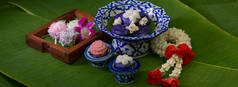 一些泰国传统甜点的近景，这些甜点用瓷器和木制托盘装饰在香蕉叶背景上的花环茉莉花