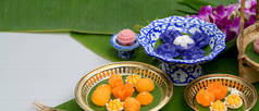 一些泰国传统甜点的近景，这些甜点放在瓷盘上，黄铜盘上有花和香蕉叶装饰在白桌上