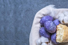 点画紫鸡蛋与自制面包。色彩艳丽的复活节彩蛋绿色帆布袋上的彩蛋是紫色的，上面有羽毛和棉铃。灰色背景上的紫色卵子。东海岸快乐