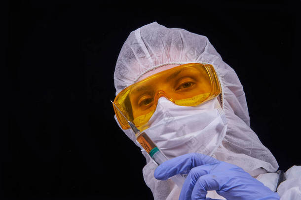 防止感染。医生身穿防护服，戴着防护眼镜，戴着防护面具，手里拿着注射器，手里拿着疫苗。在黑色背景上被隔离