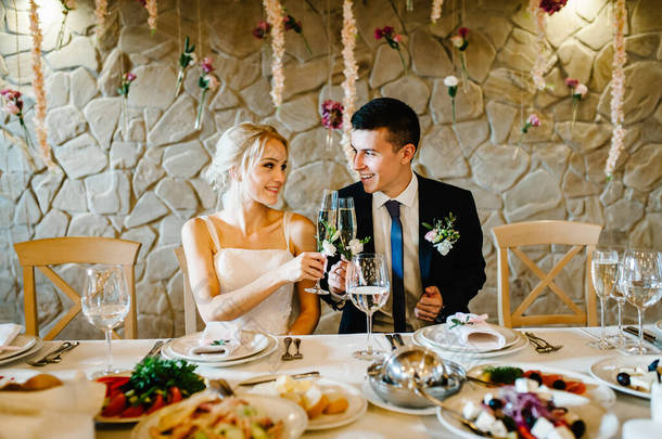 新婚夫妇举着香槟酒在屋里举杯敬酒。用鲜花和绿色装饰的婚礼仪式。餐厅宴会厅餐桌旁的<strong>新郎新娘</strong>.