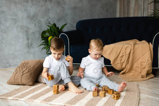 两个男孩兄弟在地板上用木制立方体块在编织的地毯上贴着地板。内饰针织风格:枕头、格子布、地毯.温暖和舒适.