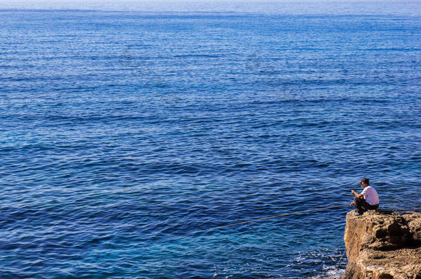 一位身份不明的老渔夫在海边的石头上，背景是蓝水蓝天- -抄袭空间 