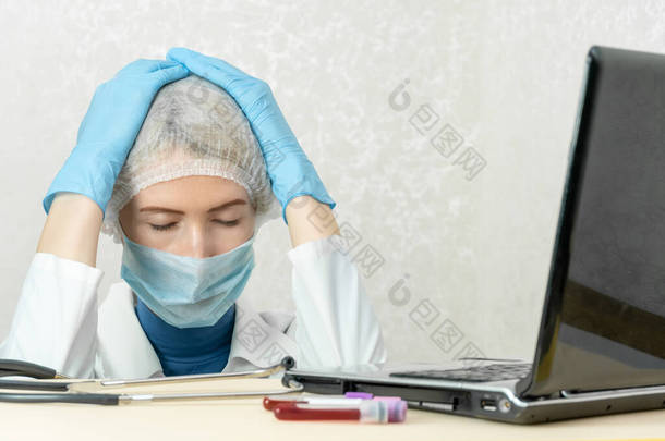 身着医疗服、戴着医疗面罩和帽子的女医生<strong>坐</strong>在笔记本电脑旁边，闭着眼睛，<strong>双手</strong>抱头。医学的概念