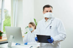 一个戴着假发的外科医生拿着一个黑色的剪贴板在医院里等着。一位女医生坐在房间角落里的台式计算机前. 