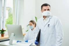 一个戴着假发的外科医生拿着一个黑色的剪贴板，在医院等病人。一位女医生坐在房间角落里的台式计算机前.