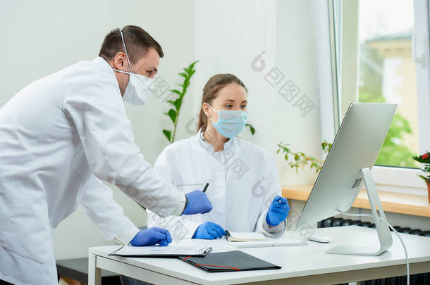 一位外科医生和一位戴口罩的放射科医生在台式计算机上讨论一位病人的X<strong>光</strong>照片。一位女医生坐在电脑前，一位治疗师呆在附近.