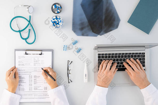 两名临床医生双手平整地做着医疗笔记，并用笔记本<strong>电脑打字</strong>，周围都是X光图像、药丸、听诊器等