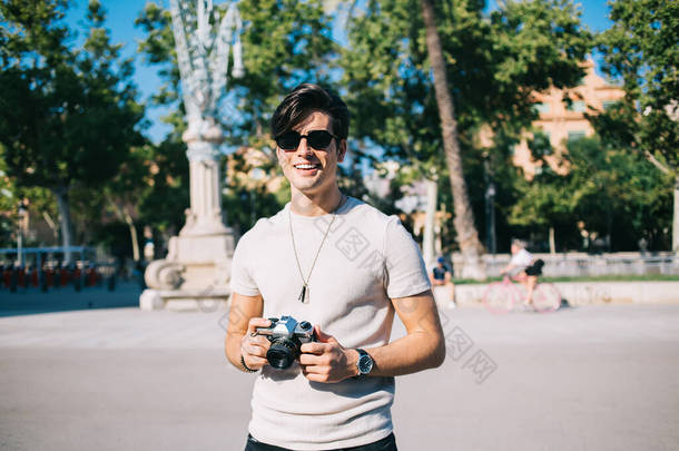 身着休闲装，戴着时髦太阳镜，拿着老式<strong>摄影</strong>相机站在<strong>城市街道</strong>上，一边欣赏巴塞罗那的散步，一边笑着
