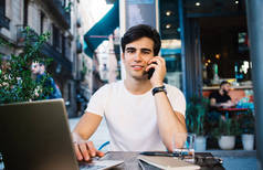 年轻英俊的男子穿着休闲装，坐在巴塞罗那市的咖啡店里，一边用笔记本电脑一边用智能手机聊天，一边看着相机