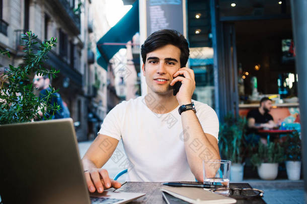 年轻英俊的男子穿着休闲装，坐在巴塞罗那市的<strong>咖啡店</strong>里，一边用笔记本电脑一边用智能手机聊天，一边看着相机