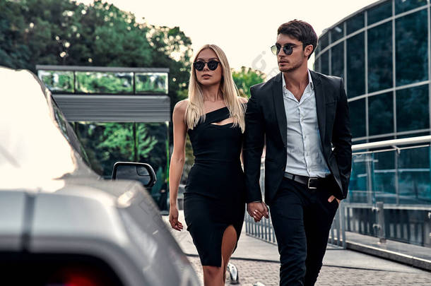 漂亮的年轻夫妇在跑车附近。迷人的金发美女穿着典雅的黑色衣服，英俊的商人穿着<strong>西服</strong>走在现代商业中心前.