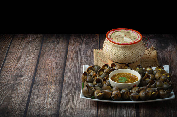 将淡水蜗牛或苹果蜗牛制成泰国菜，配上泰国菜辣辣椒酱和粘稠的饭篮放在木桌上