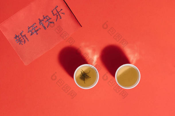 两杯中式茶杯，红面上有红茶和水蒸气。用粤语写上<strong>新年</strong>快乐字样的红包.