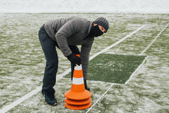 冬季，一名戴着医疗面罩的男子在体育场安排交通圆锥。蒙面男子设置交通圆锥以保持社会距离. 