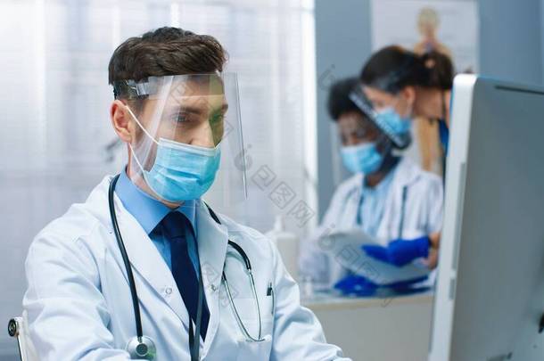 <strong>年</strong>轻英俊的白种人男医生头戴<strong>口罩</strong>坐在橱柜里的近照，在医疗中心用计算机进行检疫打字和检查医生的概念时进行面罩工作