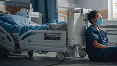 医院特护验尸官病房：悲伤、疲惫的黑人护士头戴面罩坐在地板上的画像，对所有大流行病患者表示歉意。勇敢无畏的英雄医护人员拯救生命