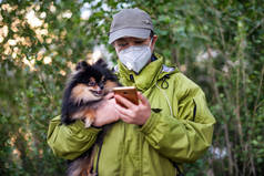 在COVID-19森林里，一个戴着口罩和带着狗散步的女孩在用智能手机拨号.
