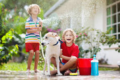 孩子们在夏天的花园里洗狗.水管和洒水的乐趣为孩子。百花盛开的后院,孩子们在户外的庭院里洗小狗.孩子们玩。带着宠物的孩子家庭浴犬。动物护理.