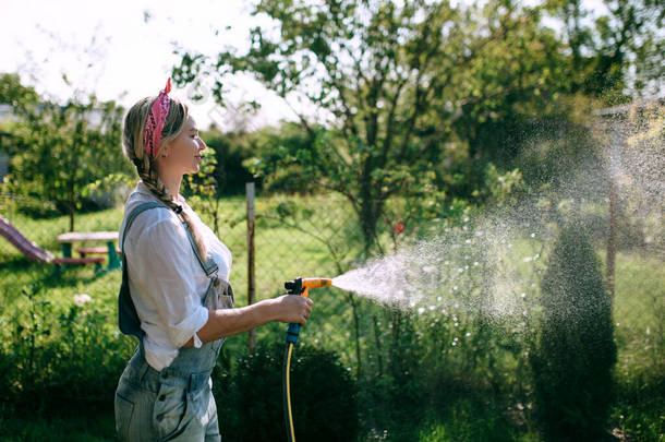 一个穿着白衬衫的年轻女人，穿着牛仔工作服在<strong>草坪</strong>上<strong>浇水</strong>。园艺概念和景观设计