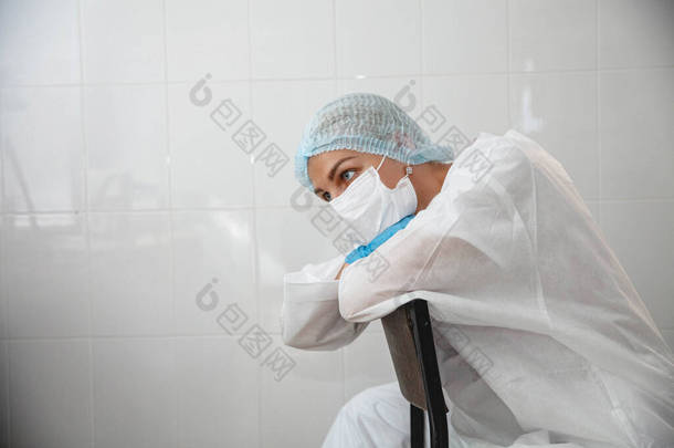 一位身穿防护服、头戴帽子、戴着面具、戴着手套的年轻女医生疲惫不堪地坐在医务室的椅子上