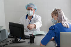 一名身穿白衣、戴着面具和帽子的医生与一名病人坐在办公室的桌子旁，并在显示器上显示检查结果。协商招待会