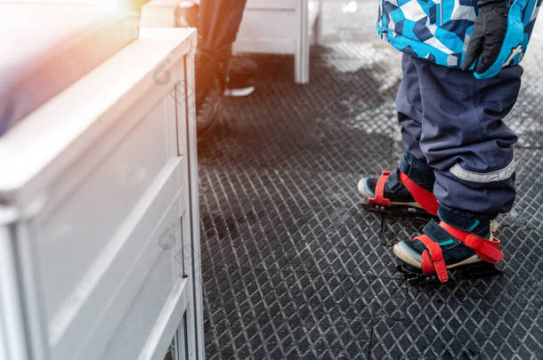 在溜冰场更衣室里，穿着蓝色塑料租借溜冰鞋站在防滑橡胶软垫上的小孩腿的特写画面。健康儿童康乐活动