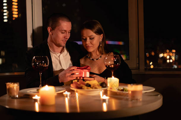 情人节这天晚上烛光下的约会，两人共进浪漫晚餐，男人在餐桌前送给女孩一份结婚周年礼物，男女关系，窗户背景，情人节的庆祝理念