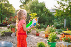 Happy girl in red dress putting out tongue shooting and splashing water with squirt gun among garden