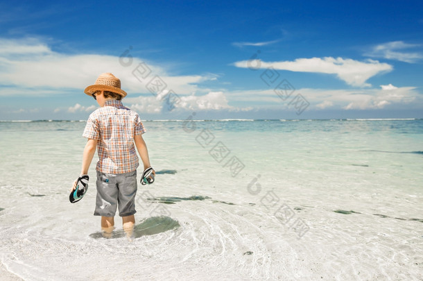 Happy young boy staying in the ocean water on beautiful beach wearing hat and sunglasses.