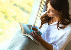 家里的年轻女人坐在窗边，在客厅里看书，喝咖啡或茶，放松身心