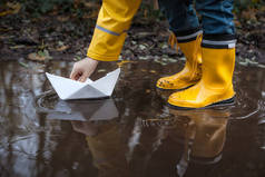 孩子与黄色的雨靴子和一个小的白色纸船/船: 在水坑中玩, 想象他的冒险