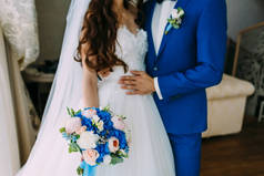 穿着白色礼服的新娘和穿着蓝色燕尾服的新郎正站在窗边, 捧着一束结婚花束。.