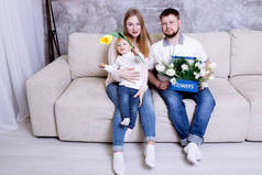 母亲、父亲和女儿坐在家里, 手里拿着鲜花。一家人在家里笑着玩。母亲节庆祝, 礼物, 郁金香