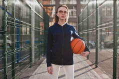 体育女孩自信在一个阳光明媚的夜晚站在篮球场上, 手里拿着篮球