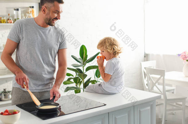 男人在煎锅里做煎饼, 而他的儿子坐在厨房的桌面上