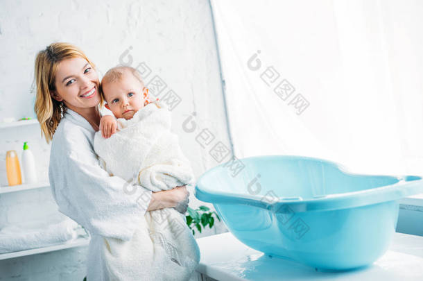 微笑的母亲在浴袍携带可爱的孩子覆盖在毛巾附近塑料婴儿浴缸