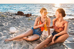 快乐的年轻女友无比的夏日饮品, 在夕阳下的沙滩上互相微笑的玻璃瓶